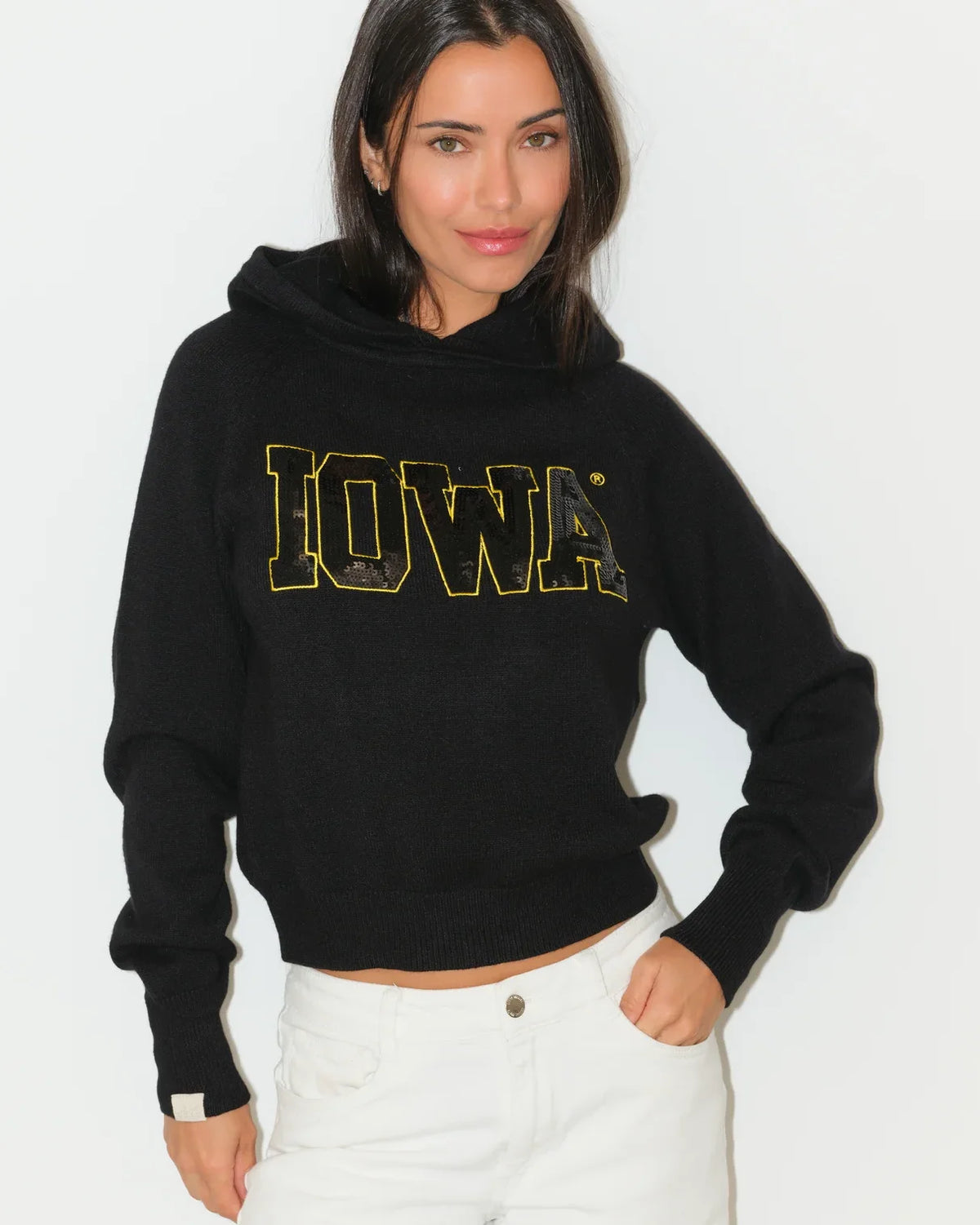 Black Iowa Hawkeyes cropped pullover hoodie featuring bold gold sequin "IOWA" lettering across the chest, modeled with white jeans.
