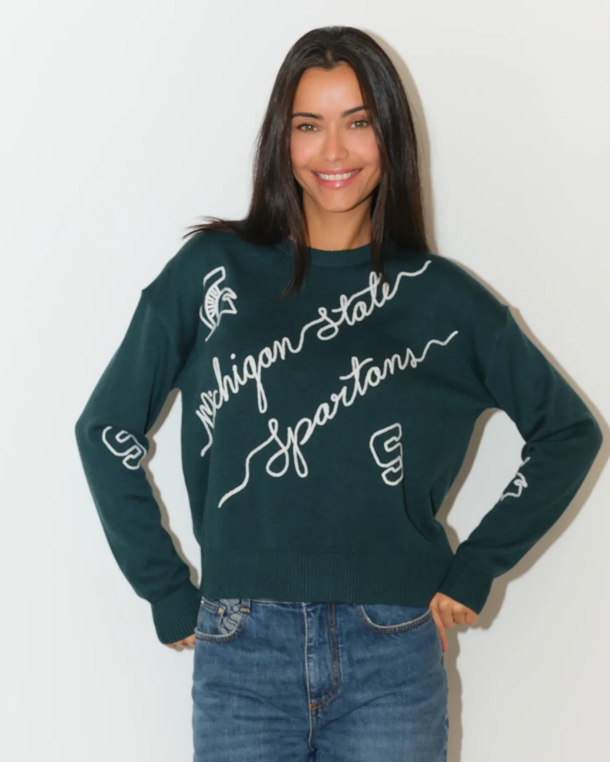 Evergreen Michigan State Spartans crew neck sweater with cursive script lettering and Spartan logo on the sleeve, paired with jeans.