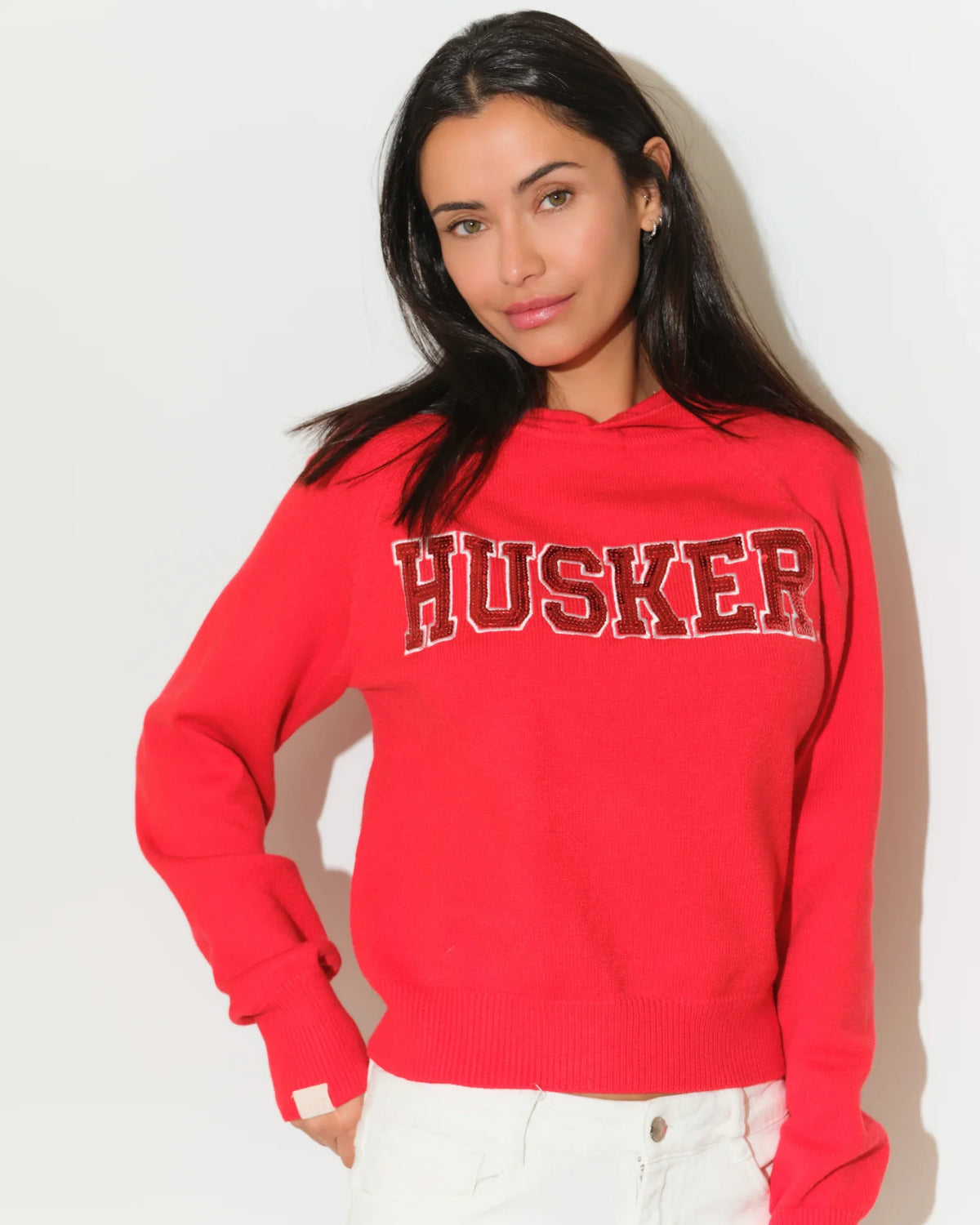 Red Nebraska Cornhuskers pullover hoodie featuring sequin "HUSKER" lettering across the chest, with a relaxed cropped fit and kangaroo pocket.