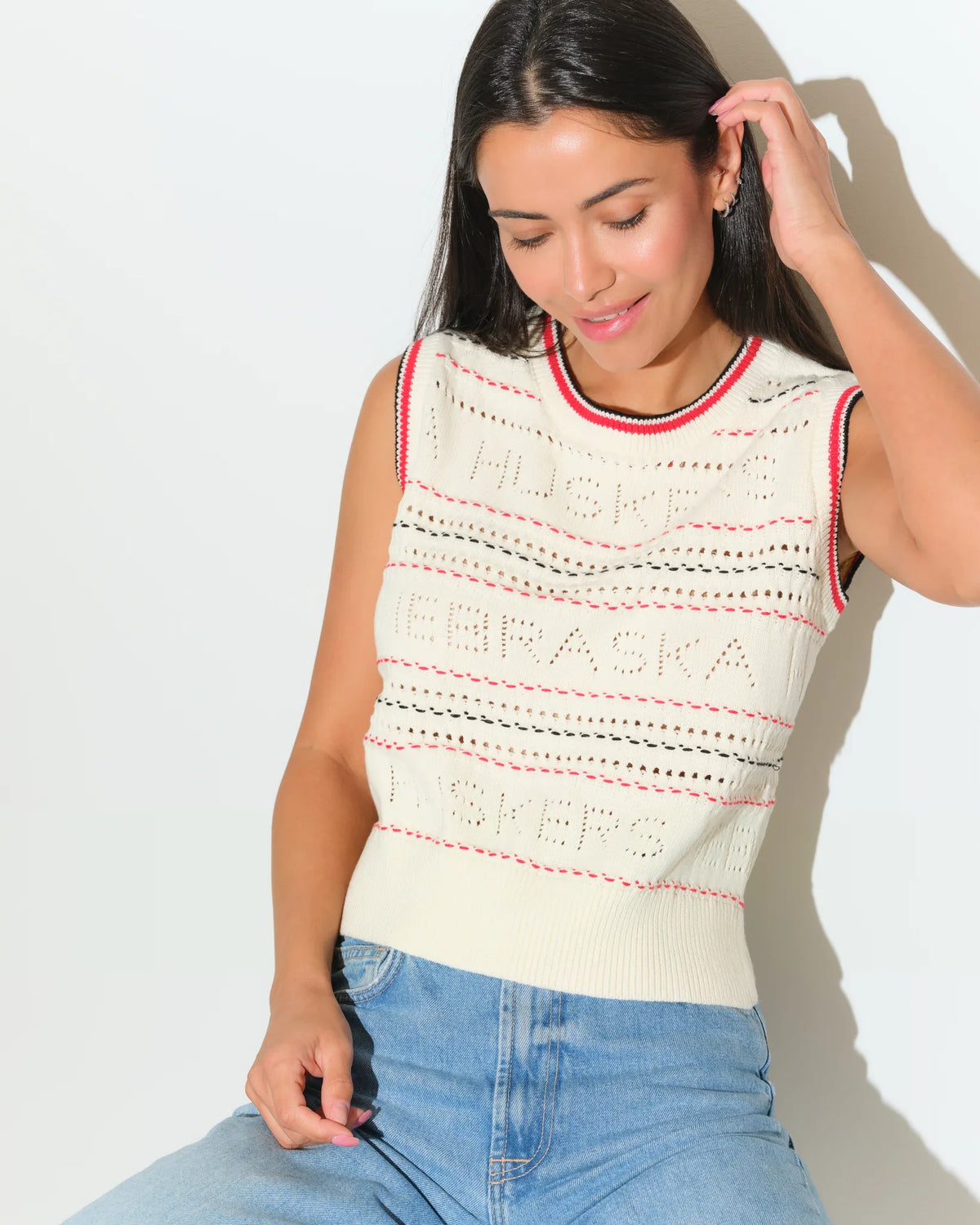 Ivory Nebraska Cornhuskers sweater vest with red trim, open-knit texture, and embroidered team lettering, styled with jeans.