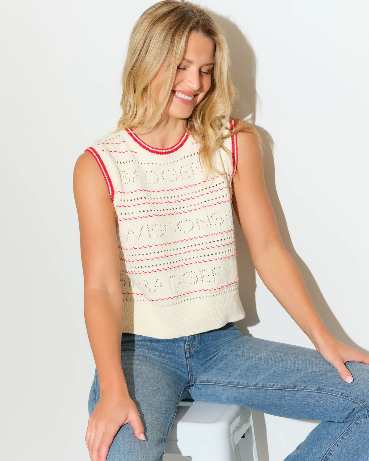 Ivory Wisconsin Badgers sweater vest with red trim accents and open-knit texture, styled with jeans.