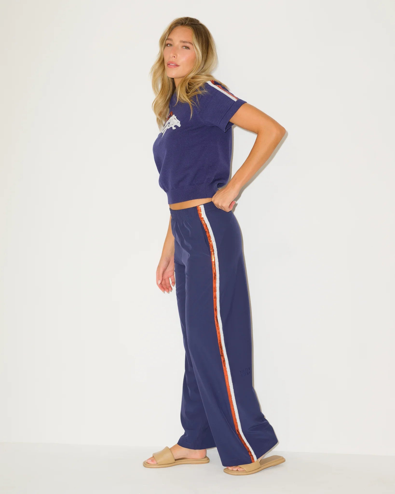 Navy KADYLUXE sequin track pants with a bold side stripe, styled with a matching Denver Broncos tee. Full-length silhouette with a relaxed, sporty fit.