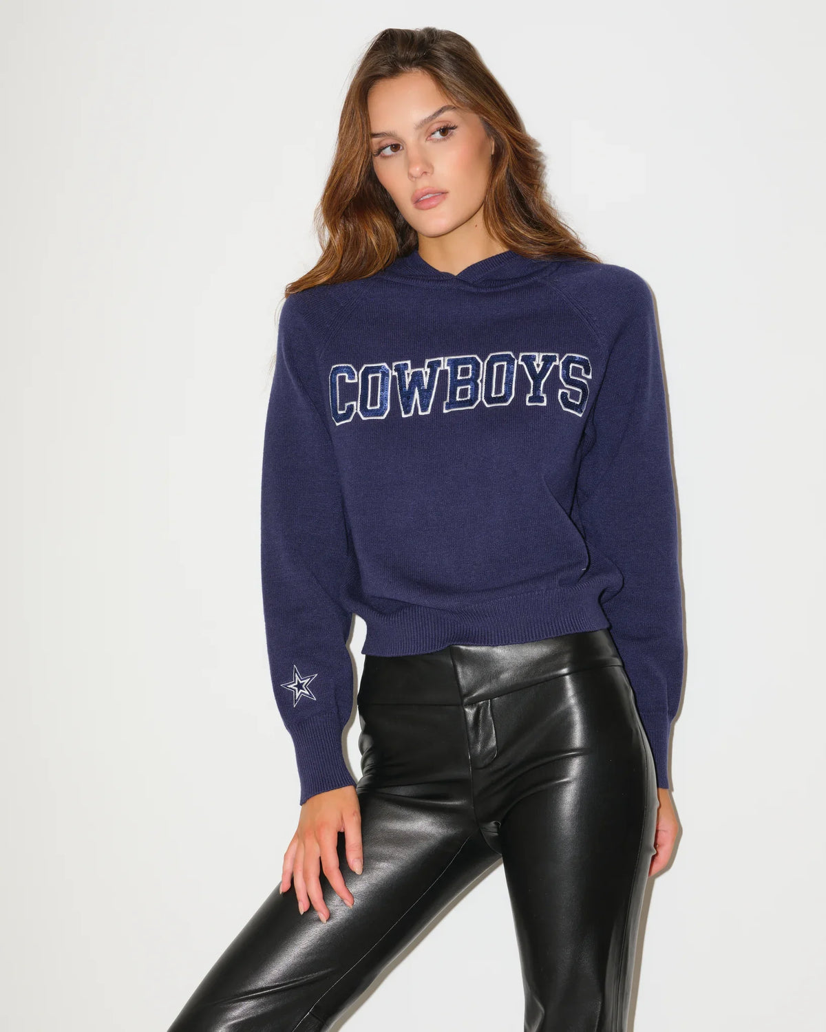 Navy Dallas Cowboys Shine Knit Hoodie with bold "COWBOYS" lettering across the chest, worn by a seated model paired with black leather pants.