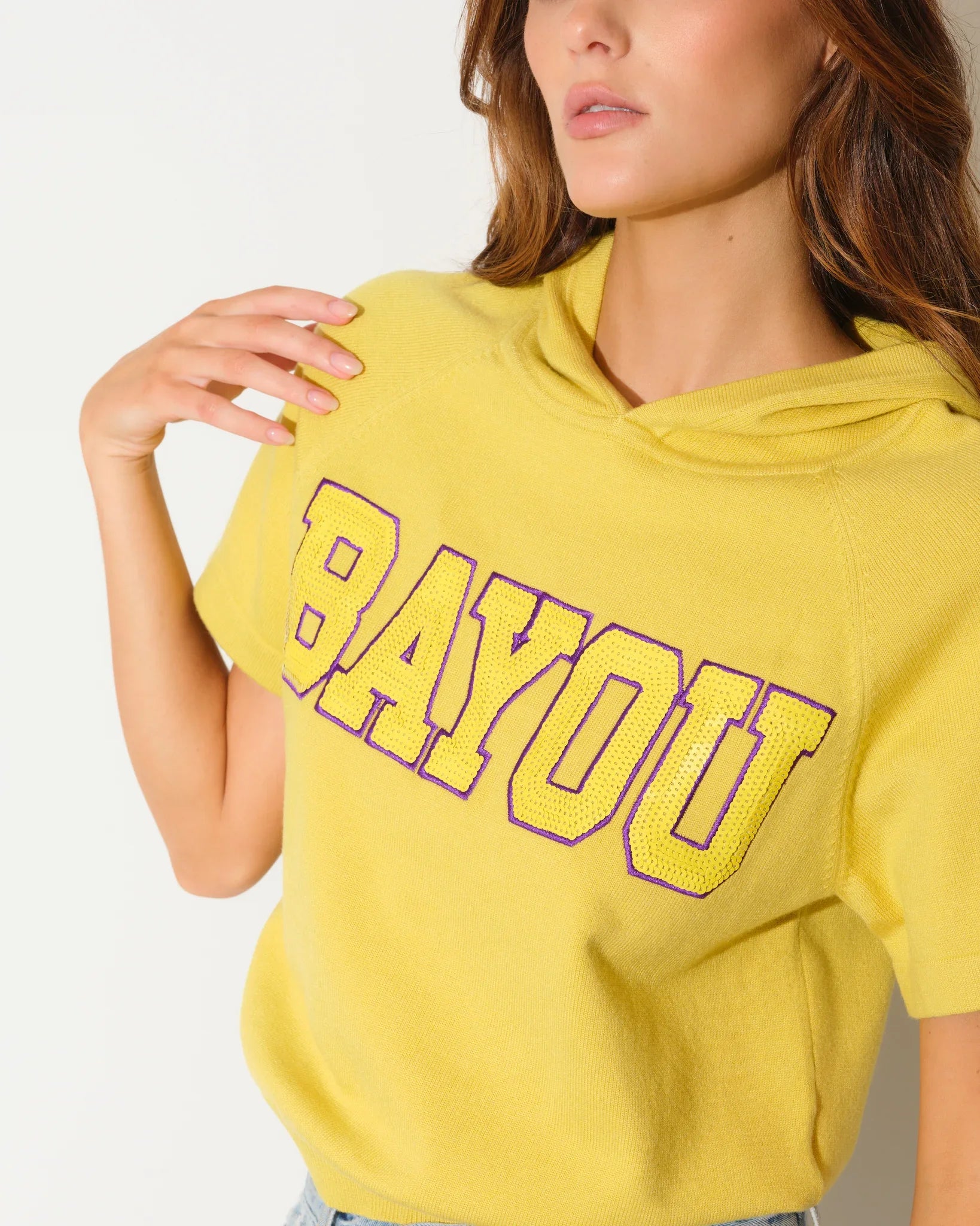 Yellow short sleeve cropped hoodie with bold "BAYOU" varsity lettering across the chest, featuring subtle sequin texture throughout the knit fabric.