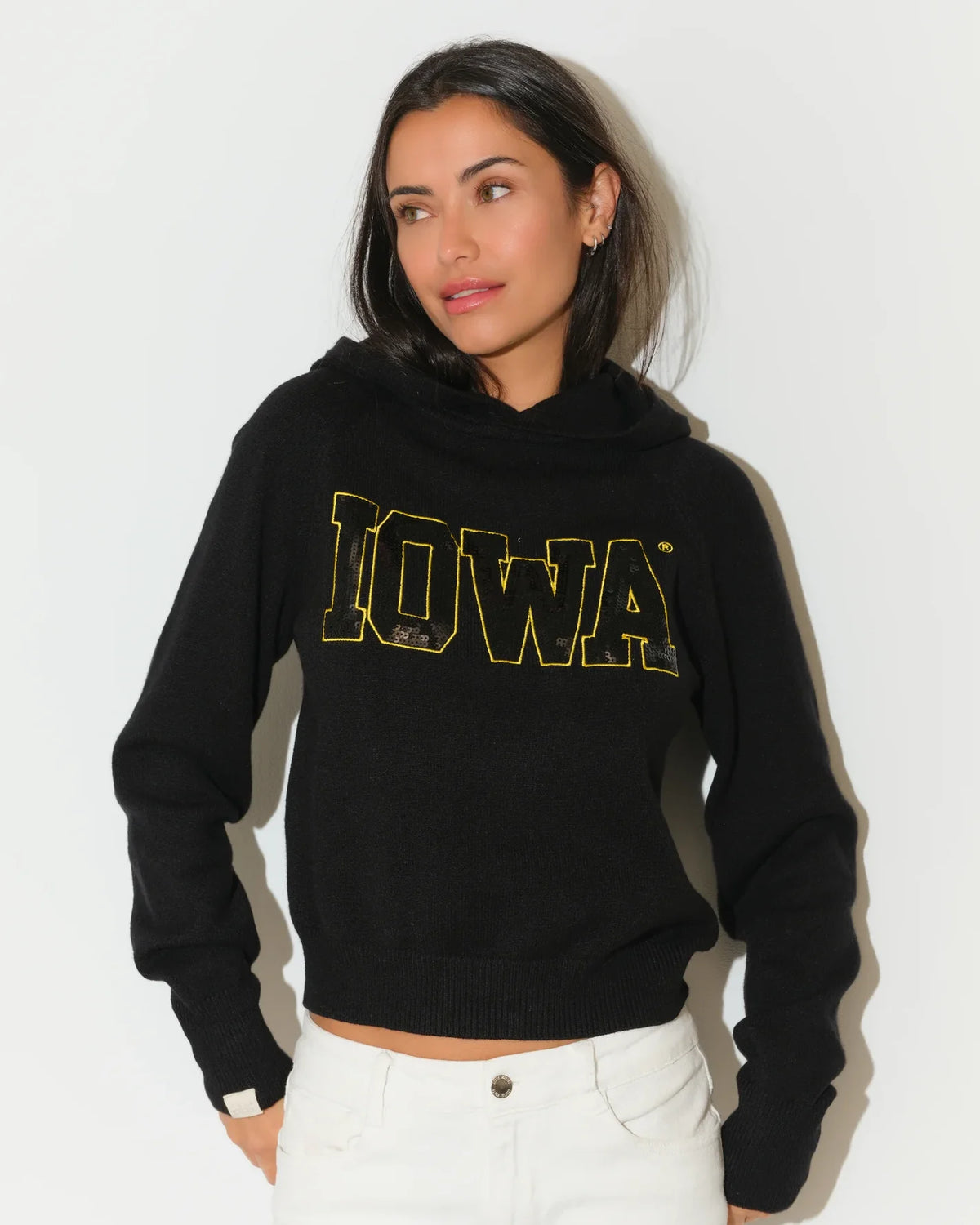 Black Iowa Hawkeyes cropped pullover hoodie featuring bold gold sequin "IOWA" lettering across the chest, modeled with white jeans.
