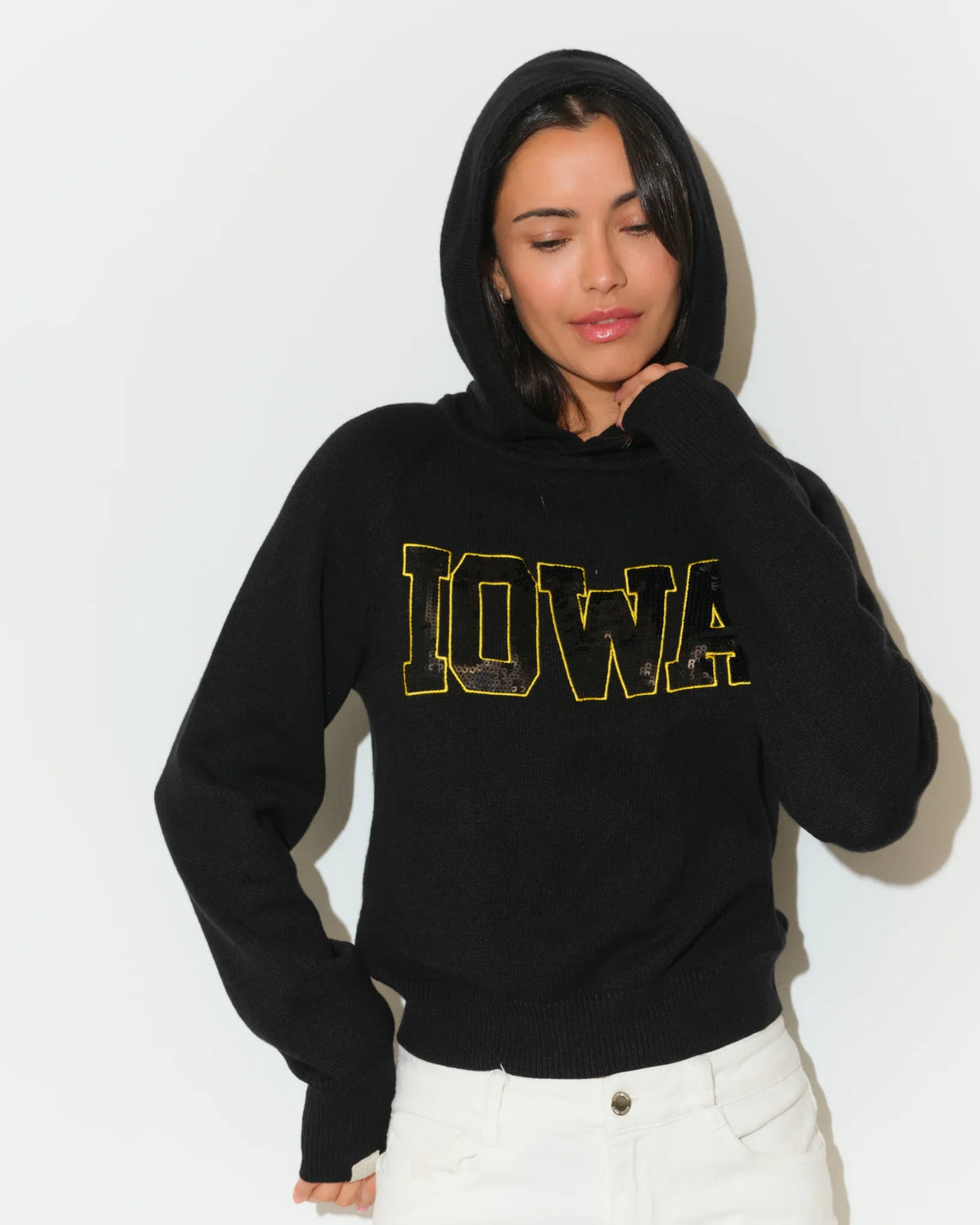Black Iowa Hawkeyes cropped pullover hoodie featuring bold gold sequin "IOWA" lettering across the chest, modeled with white jeans.