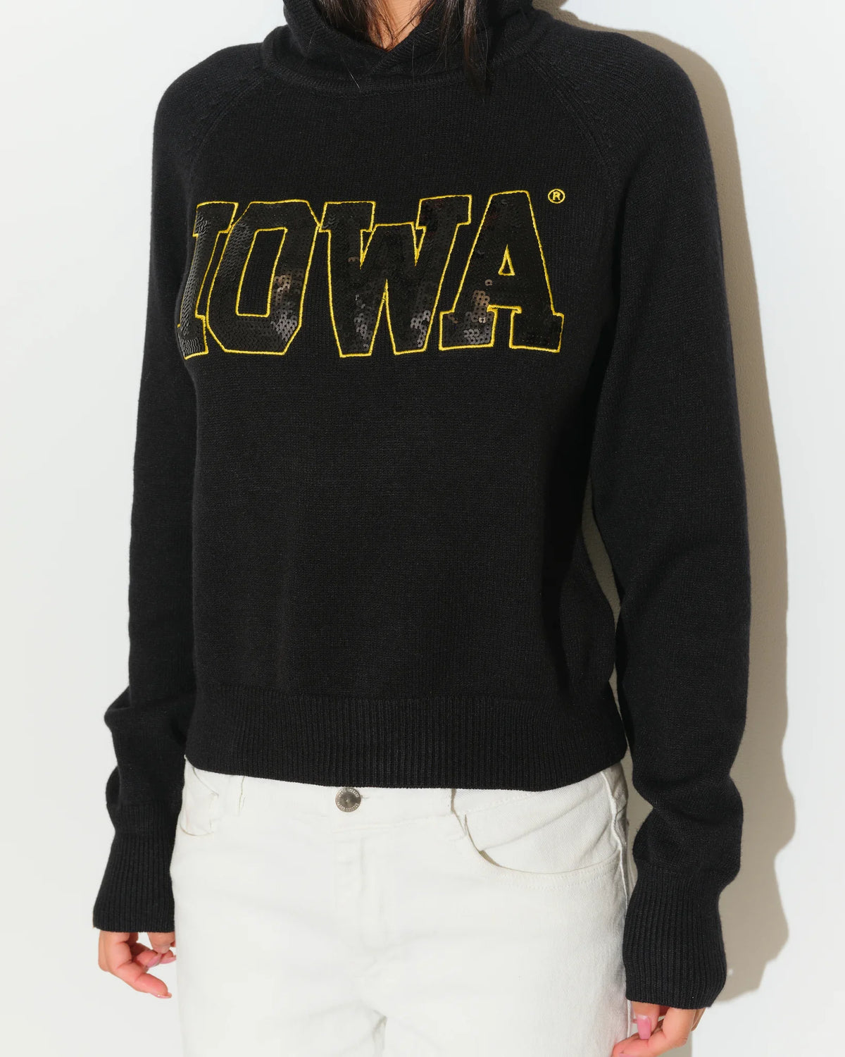 Black Iowa Hawkeyes cropped pullover hoodie featuring bold gold sequin "IOWA" lettering across the chest, modeled with white jeans.