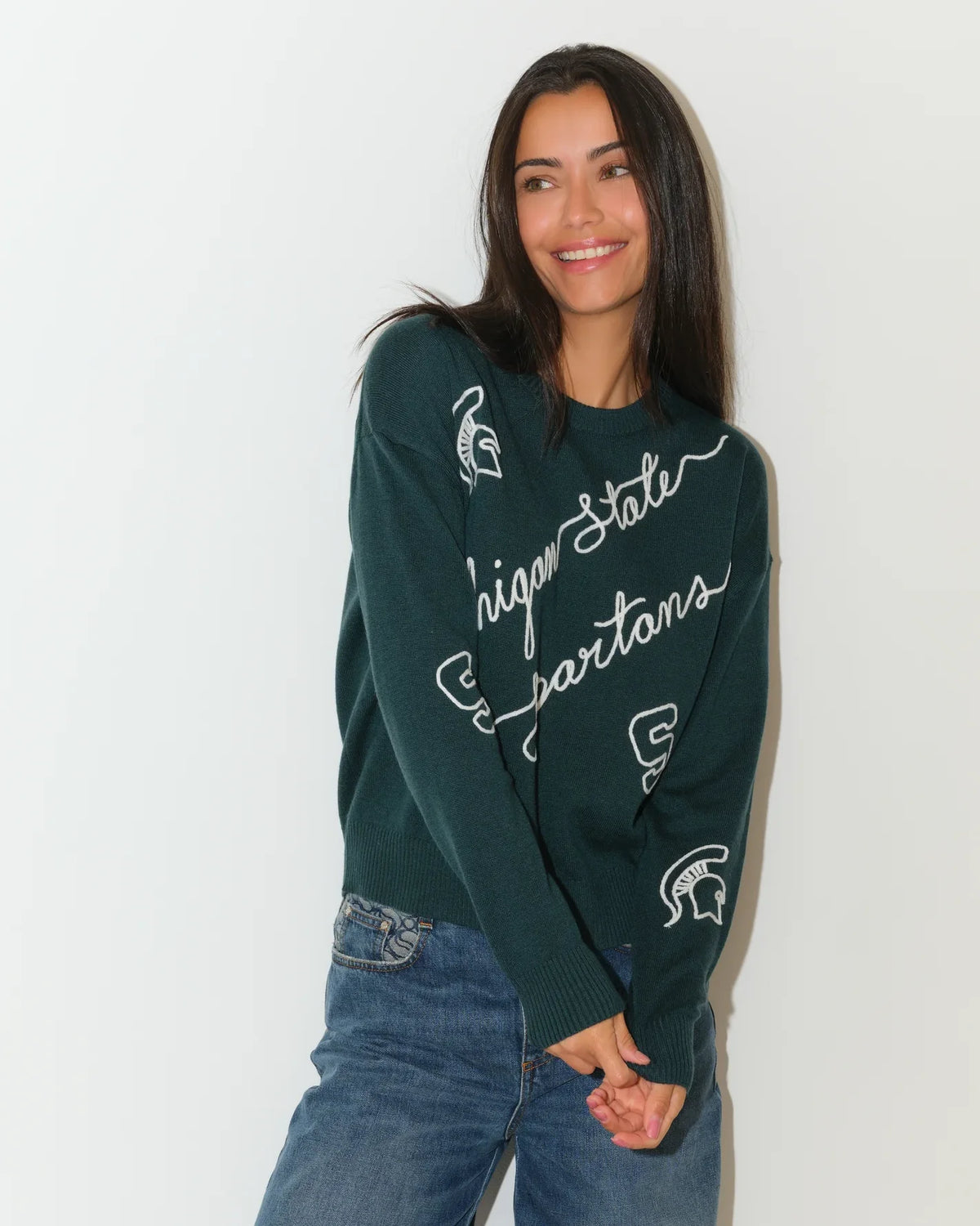 Evergreen Michigan State Spartans crew neck sweater with cursive script lettering and Spartan logo on the sleeve, paired with jeans.