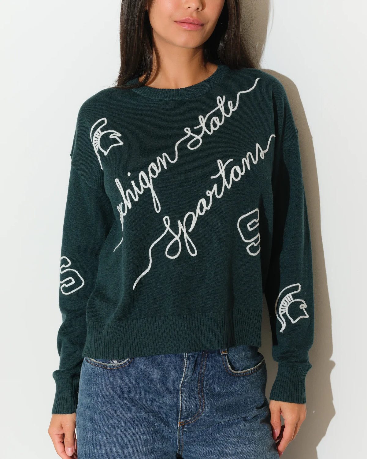 Evergreen Michigan State Spartans crew neck sweater with cursive script lettering and Spartan logo on the sleeve, paired with jeans.