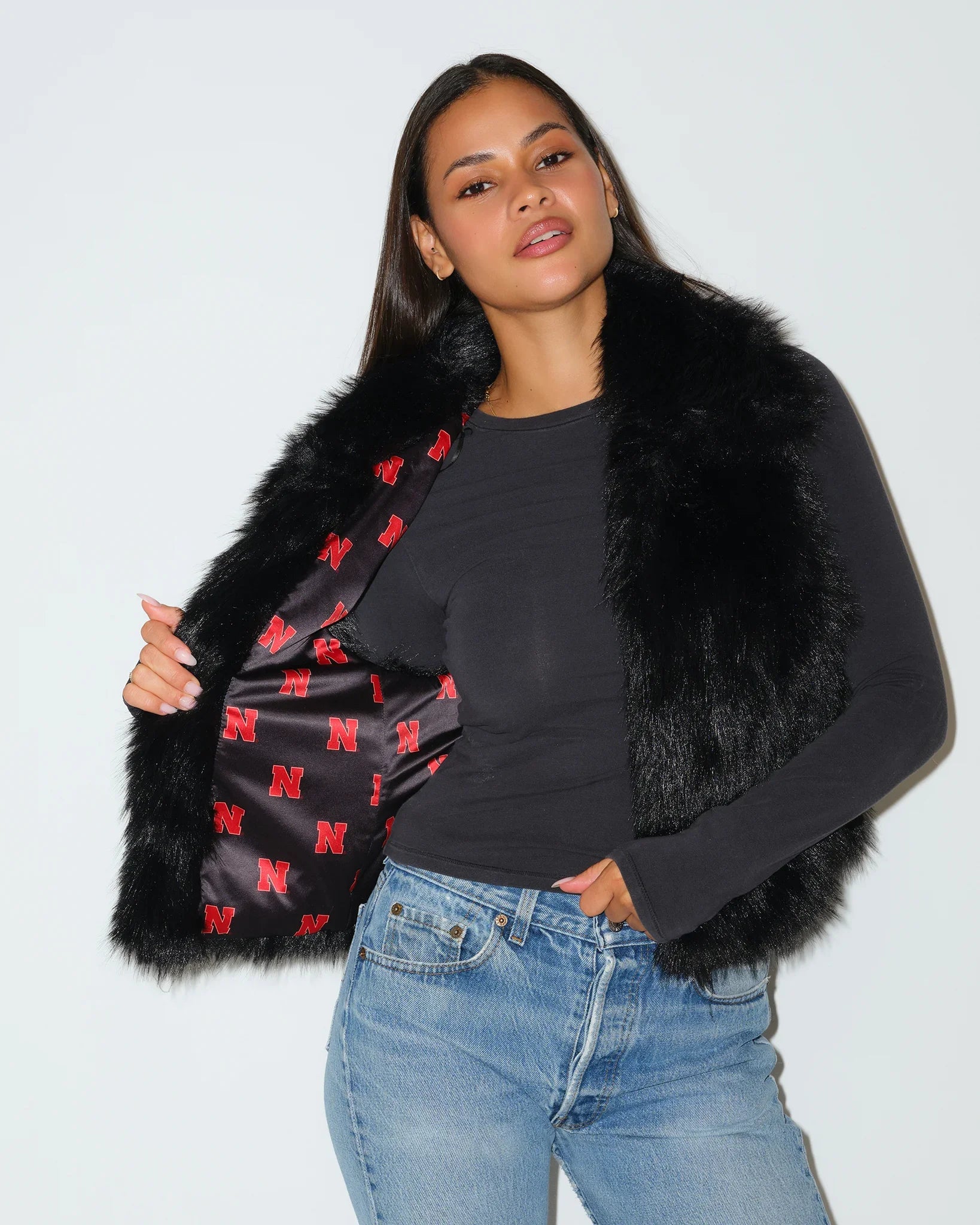 Black Nebraska Cornhuskers faux fur vest with red plaid Huskers-print lining, worn open over a gray long-sleeve top with jeans.