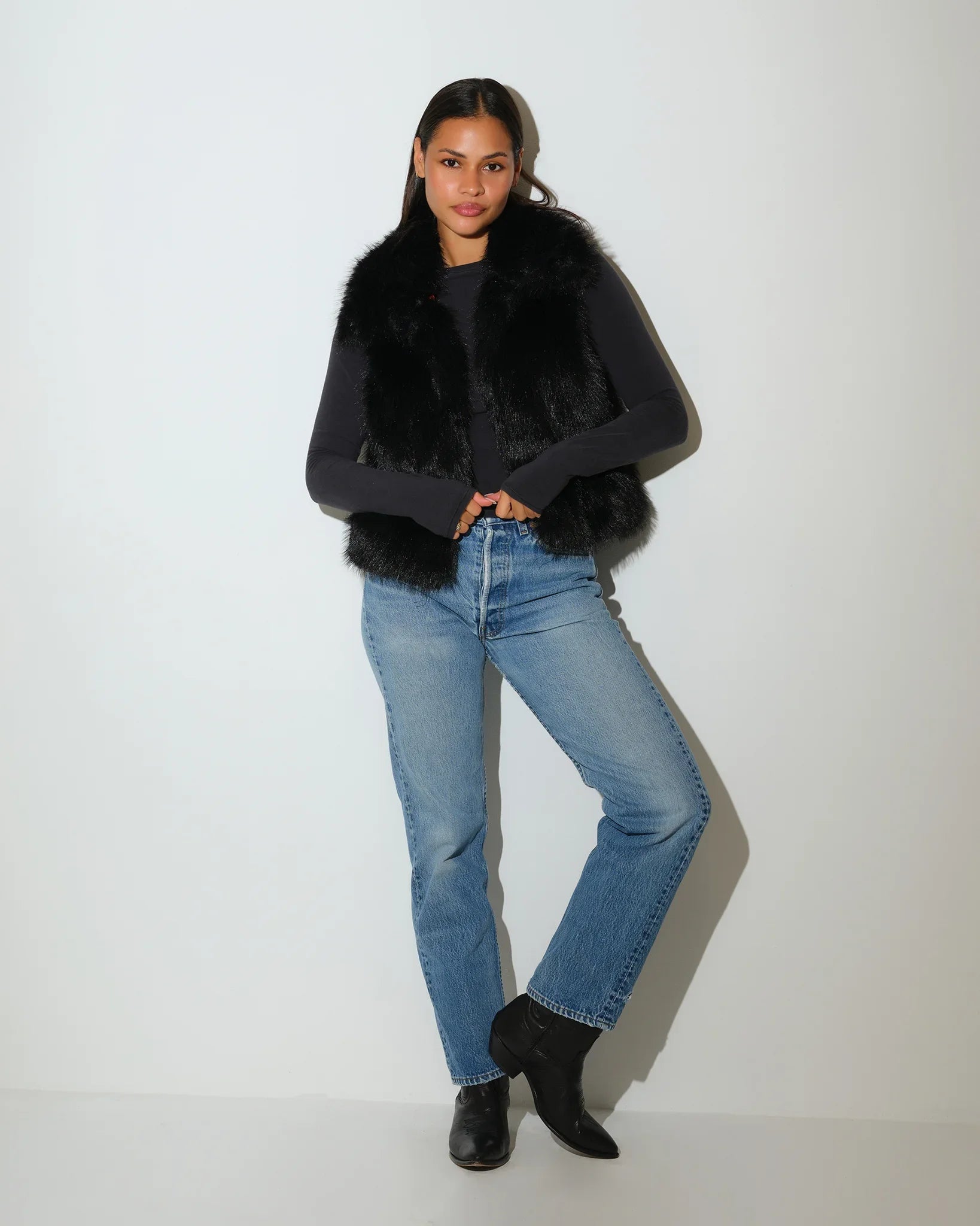 Black Nebraska Cornhuskers faux fur vest with red plaid Huskers-print lining, worn open over a gray long-sleeve top with jeans.