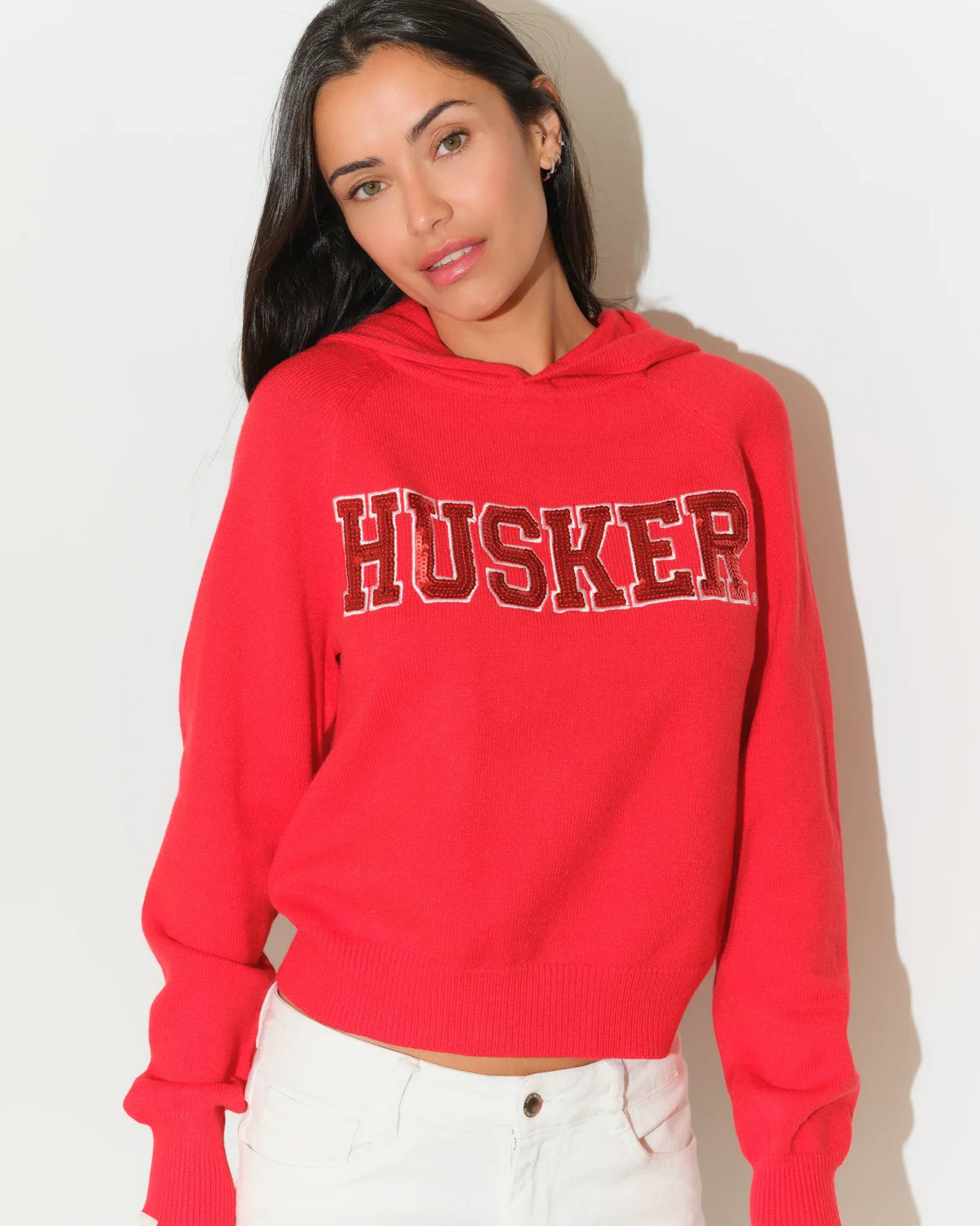 Red Nebraska Cornhuskers pullover hoodie featuring sequin "HUSKER" lettering across the chest, with a relaxed cropped fit and kangaroo pocket.