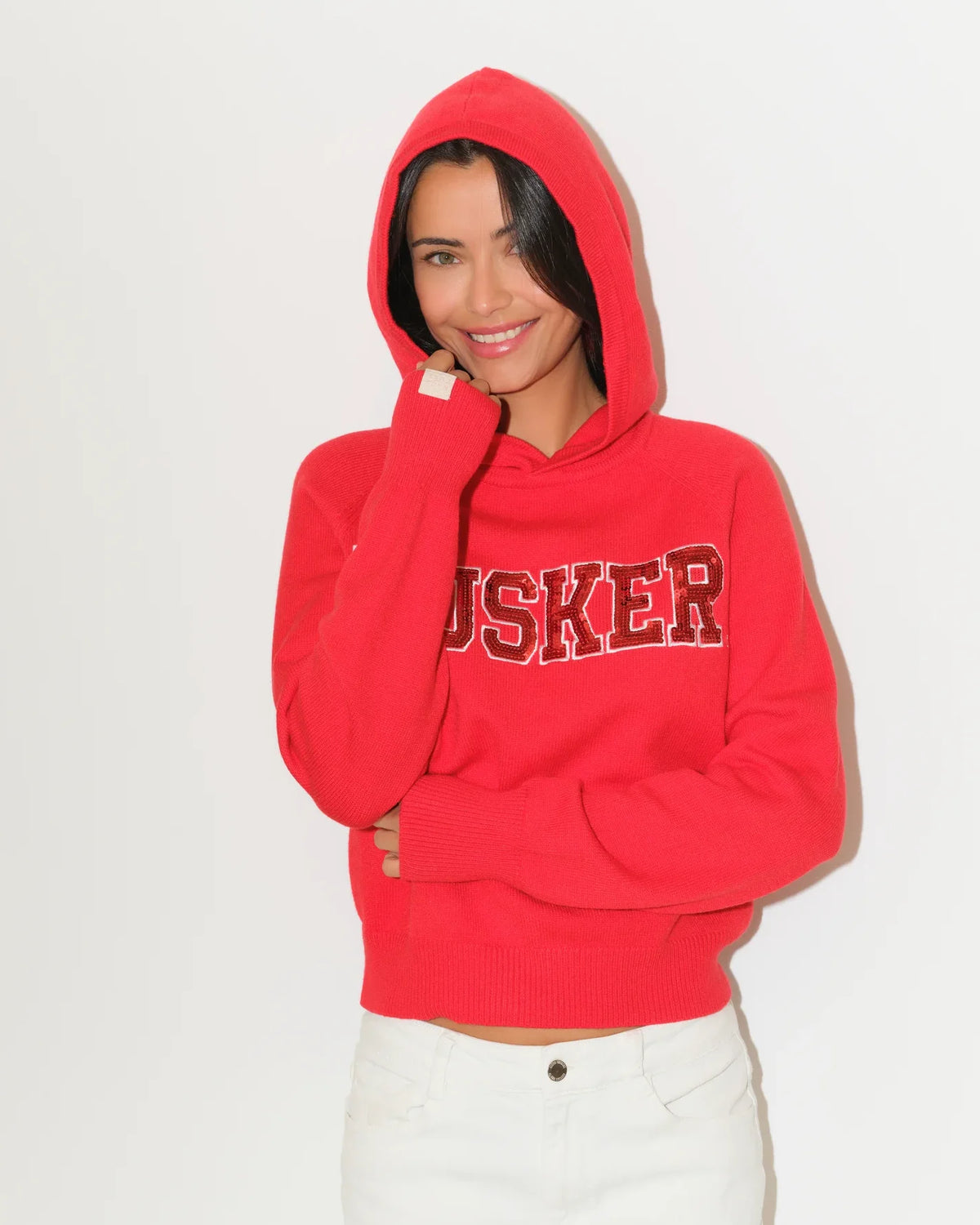 Red Nebraska Cornhuskers pullover hoodie featuring sequin "HUSKER" lettering across the chest, with a relaxed cropped fit and kangaroo pocket.