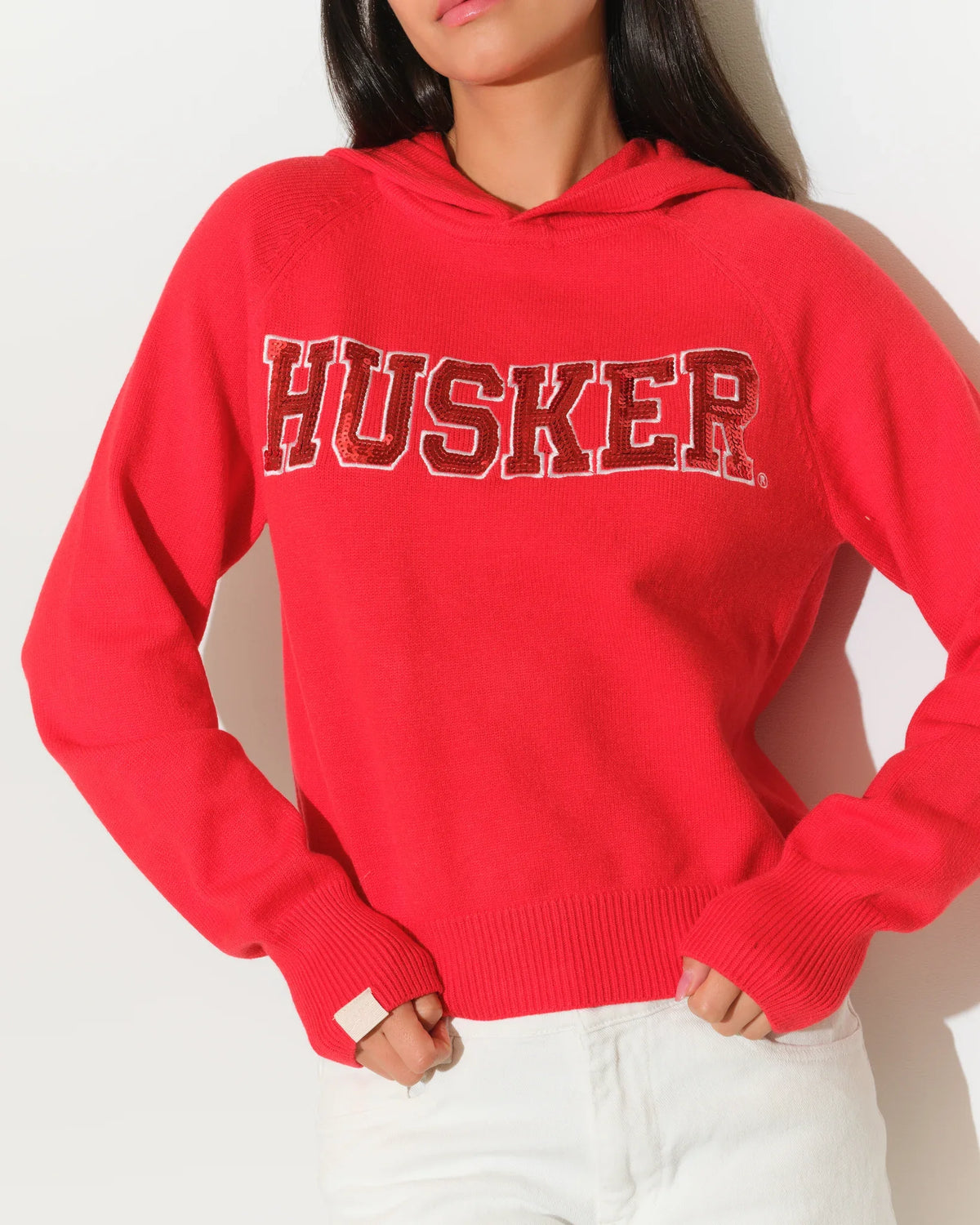 Red Nebraska Cornhuskers pullover hoodie featuring sequin "HUSKER" lettering across the chest, with a relaxed cropped fit and kangaroo pocket.
