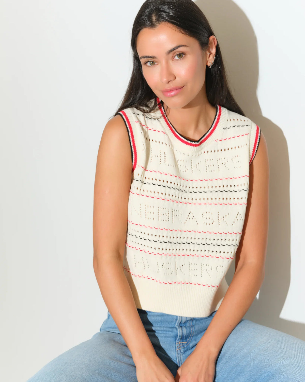Ivory Nebraska Cornhuskers sweater vest with red trim, open-knit texture, and embroidered team lettering, styled with jeans.