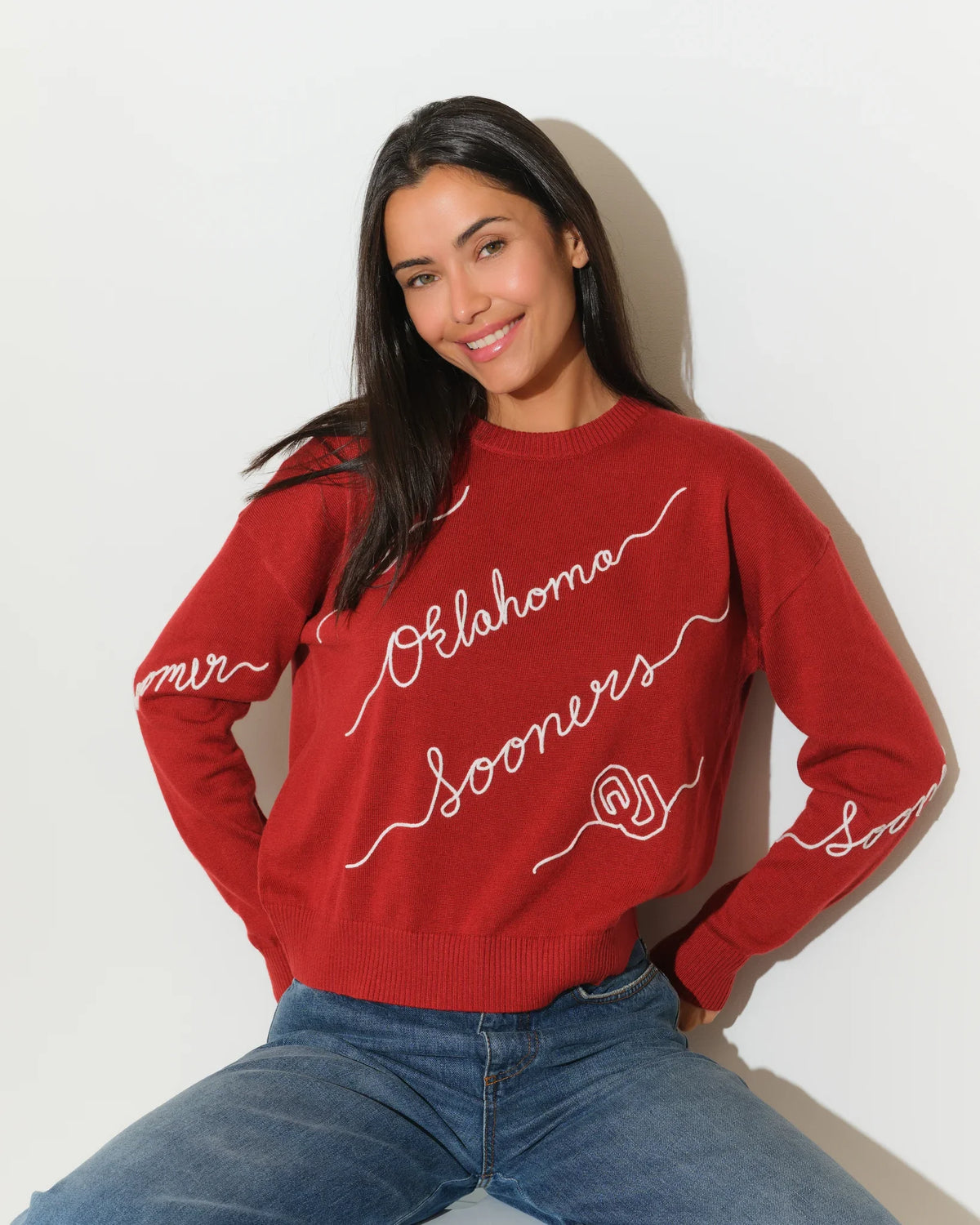 Crimson Oklahoma Sooners crew neck sweater with cursive script lettering across the front, paired with jeans.