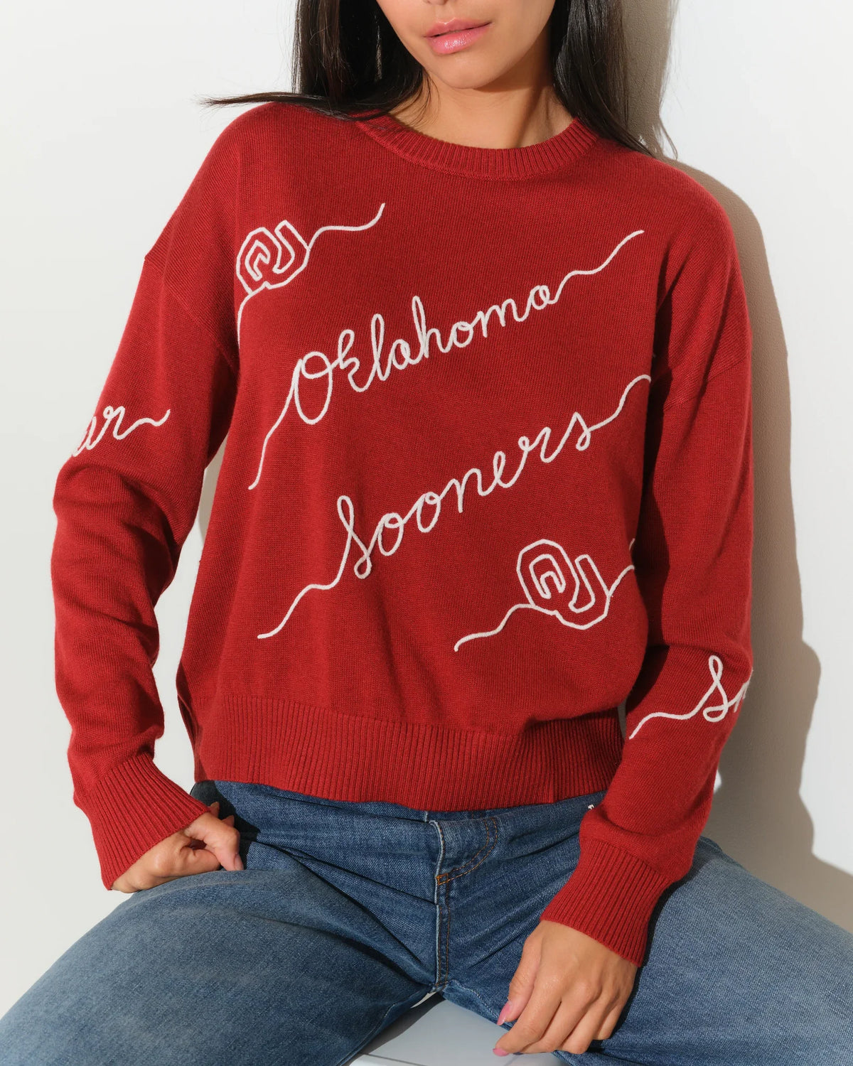 Crimson Oklahoma Sooners crew neck sweater with cursive script lettering across the front, paired with jeans.