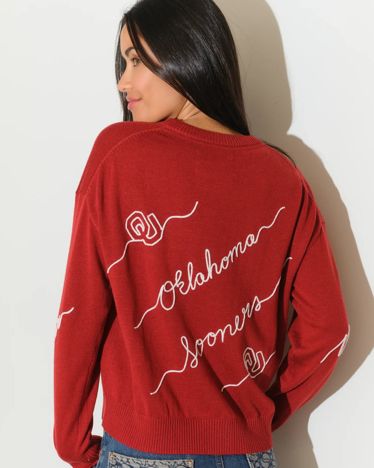 Crimson Oklahoma Sooners crew neck sweater with cursive script lettering across the front, paired with jeans.