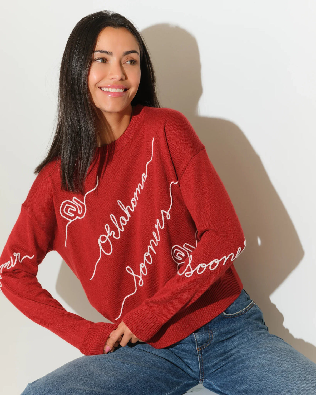 Crimson Oklahoma Sooners crew neck sweater with cursive script lettering across the front, paired with jeans.