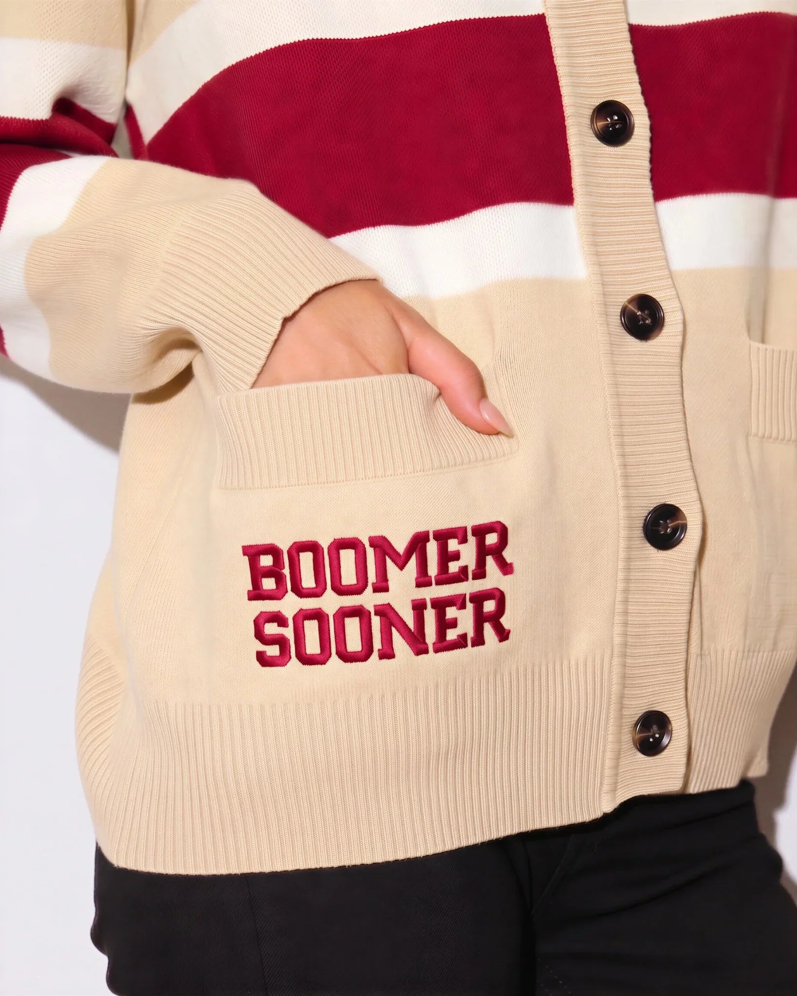 Cream Oklahoma Sooners oversized cardigan with crimson and white chest stripes, gold buttons, a helmet patch, and "Boomer Sooner" lettering on the front pocket.