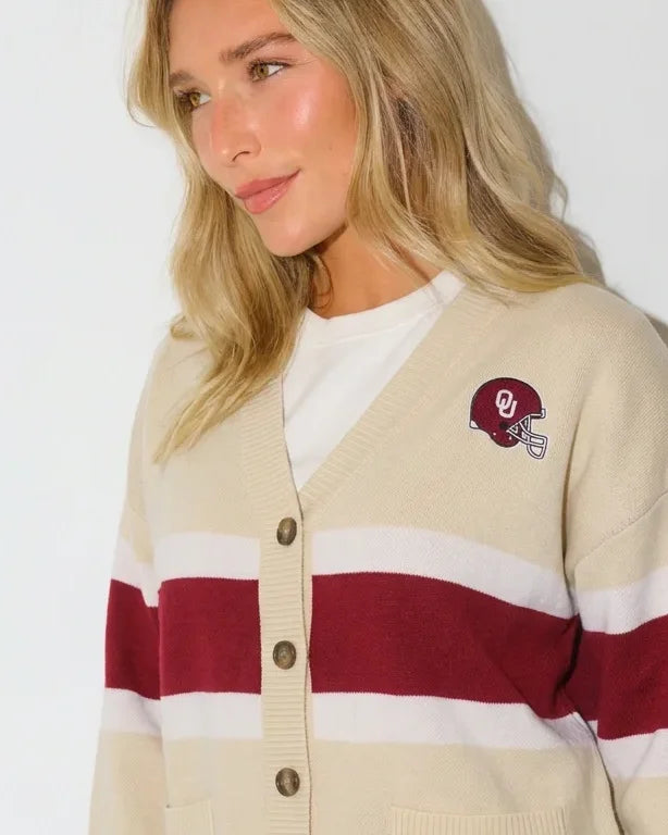Cream Oklahoma Sooners oversized cardigan with crimson and white chest stripes, gold buttons, a helmet patch, and "Boomer Sooner" lettering on the front pocket.