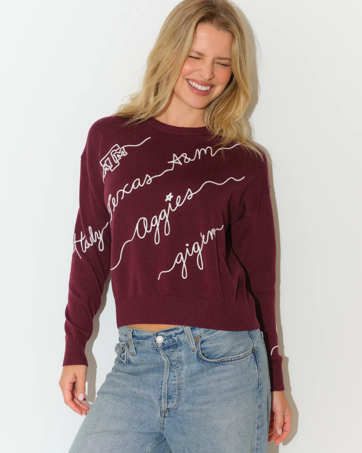 Maroon Texas A&M Aggies cropped crewneck sweater with white cursive script lettering across the front, paired with light wash jeans.
