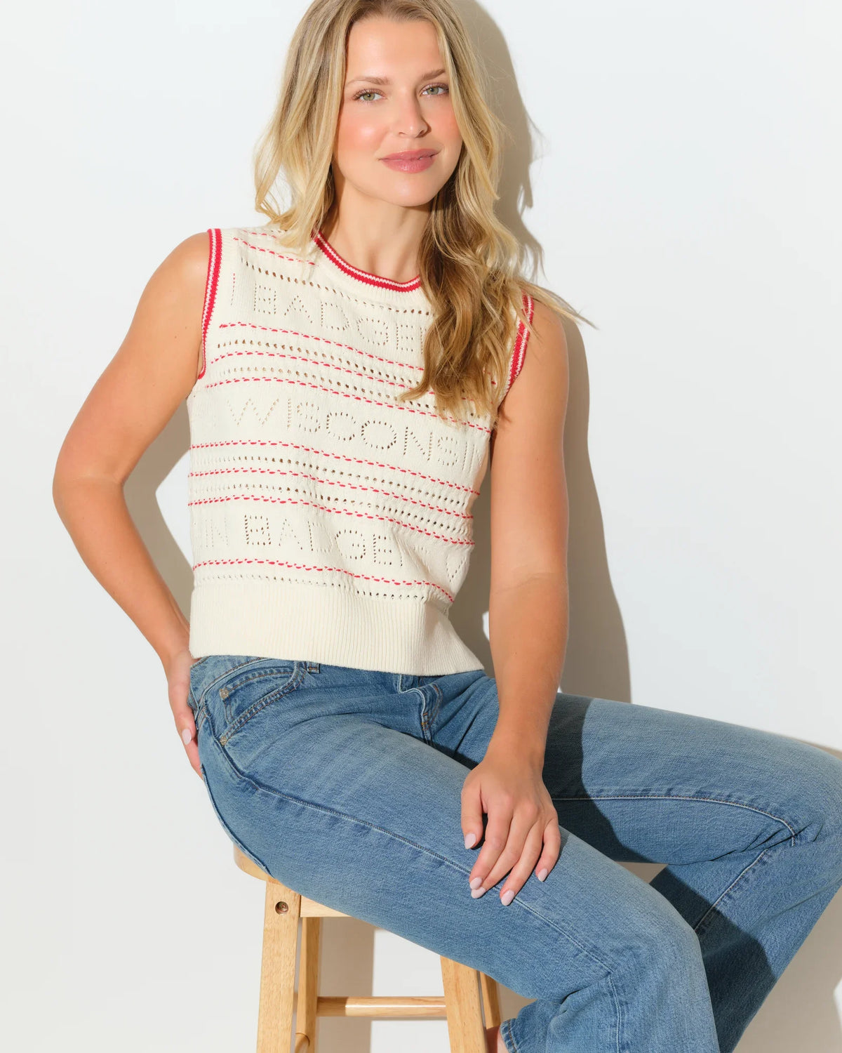Ivory Wisconsin Badgers sweater vest with red trim accents and open-knit texture, styled with jeans.