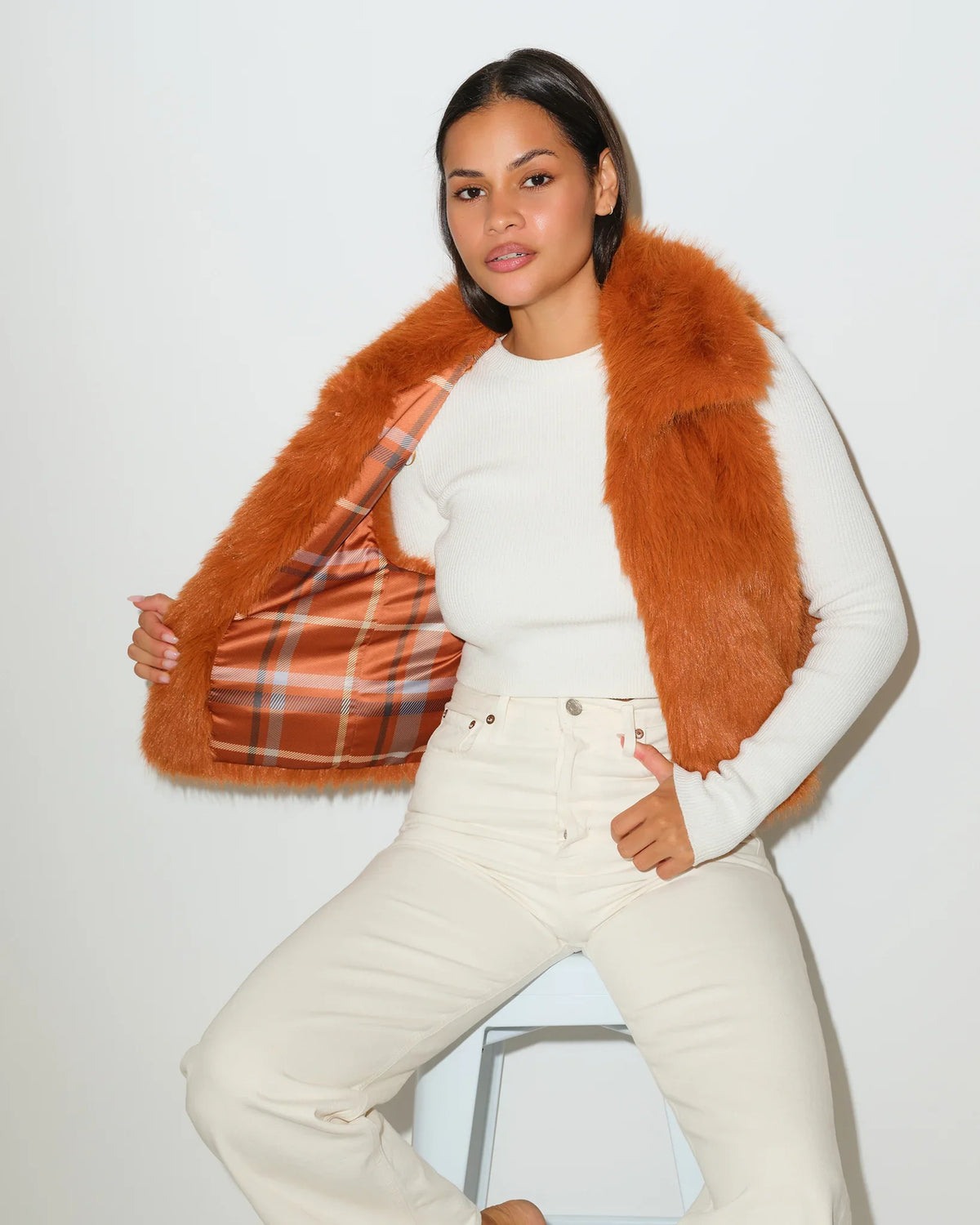 Saddle brown KADYLUXE faux fur vest with plaid lining, worn open over a white top with cream pants.