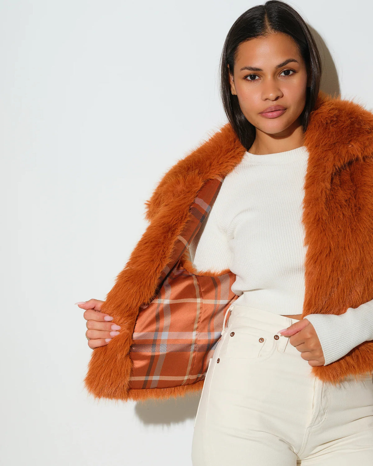 Saddle brown KADYLUXE faux fur vest with plaid lining, worn open over a white top with cream pants.