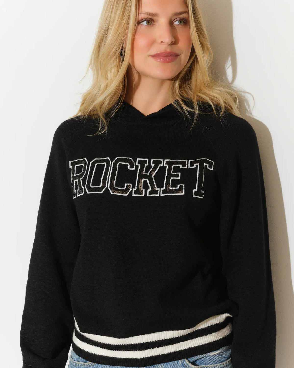 Black KADYLUXE Rocket sequin knit hoodie with bold "ROCKET" lettering and white varsity stripes at the cuffs and hem.