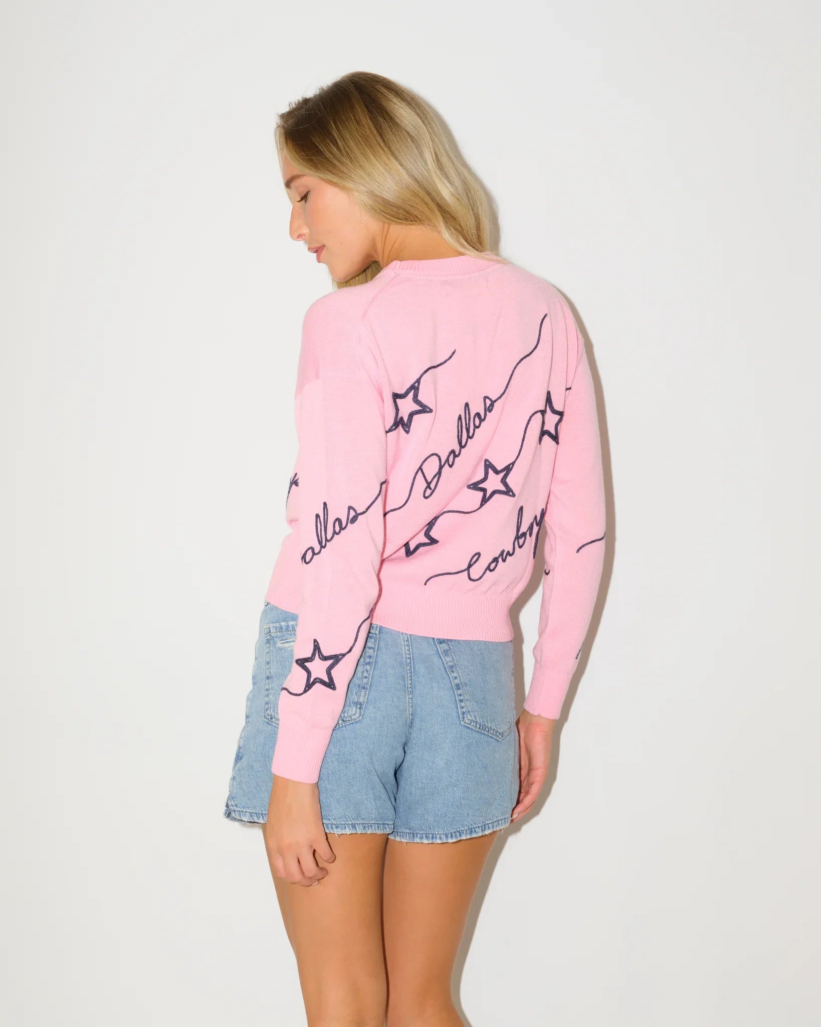 Pink Dallas Cowboys women's knit sweater with navy star pattern and script "Dallas Cowboys" lettering throughout the fabric.